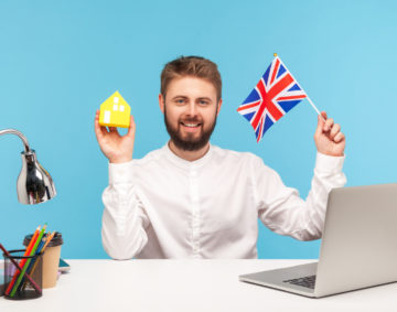 Self confident bearded man sitting at workplace with laptop, holding Great Britain flag and paper house, dreaming to buy accommodation in England. Indoor studio shot isolated on blue background.